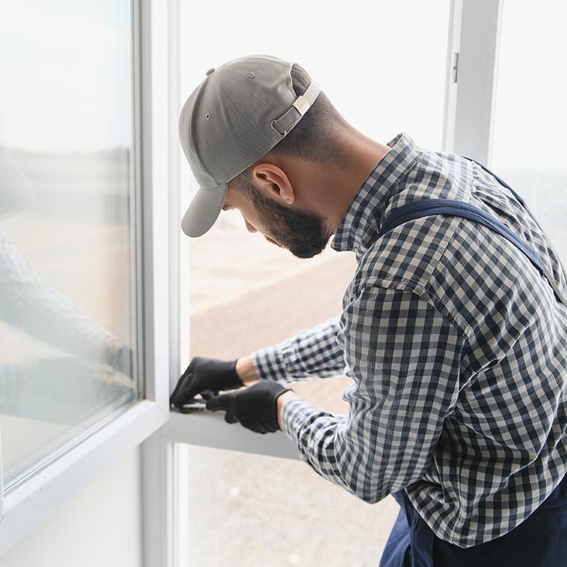 Poseur de menuiserie habillé avec une chemise à carreau et une casquette grise, qui fait un réglage sur une fenêtre PVC